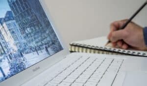A person taking notes in a notebook beside an open laptop displaying an urban scene. — Photo by mali maeder on Pexels