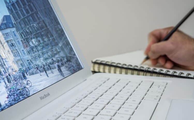 A person taking notes in a notebook beside an open laptop displaying an urban scene. — Photo by mali maeder on Pexels